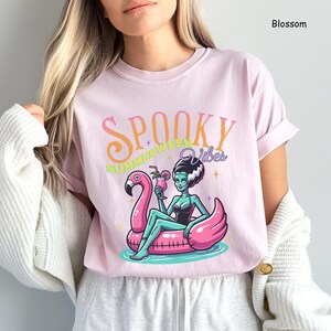 May include: A light pink t-shirt with the words "Spooky Summer Vibes" in orange and green. The graphic features a cartoon of a woman in a black swimsuit, sitting on a pink flamingo float, holding a cocktail. The shirt is a Blossom color.