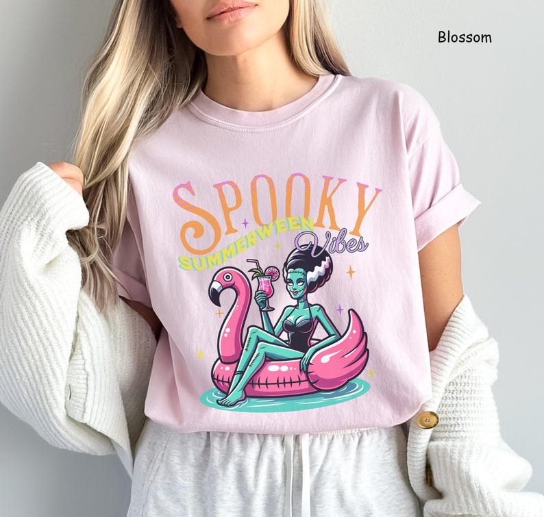 May include: A light pink t-shirt with the words "Spooky Summer Vibes" in orange and green. The graphic features a cartoon of a woman in a black swimsuit, sitting on a pink flamingo float, holding a cocktail. The shirt is a Blossom color.