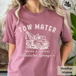 Pu&ograve; includere: T-shirt color malva melange con la scritta "TOW MATER" sopra un'illustrazione di un carro attrezzi vintage. Sotto il camion, il testo recita "TOWING & SALVAGE" e "Radiator Springs". La maglietta ha anche il testo "Magical Monday Market Original Design".