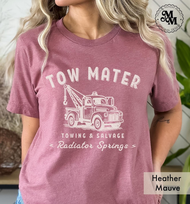 Pu&ograve; includere: T-shirt color malva melange con la scritta "TOW MATER" sopra un'illustrazione di un carro attrezzi vintage. Sotto il camion, il testo recita "TOWING & SALVAGE" e "Radiator Springs". La maglietta ha anche il testo "Magical Monday Market Original Design".
