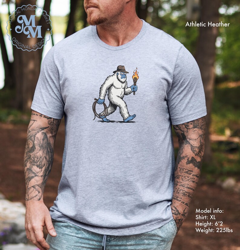 Peut inclure: T-shirt gris chin&eacute; avec un dessin d'un y&eacute;ti portant un chapeau, une torche et une corde. Le t-shirt porte le texte "Magical Monda Market Original Design" en haut &agrave; gauche. Info mod&egrave;le : Taille XL, Hauteur : 188 cm, Poids : 102 kg.