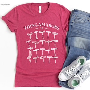 May include: A heather raspberry t-shirt with a white graphic of the text "Thingamabobs 20" and a collection of different corkscrews. The t-shirt is paired with blue jeans and gray sneakers.