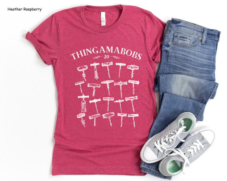 May include: A heather raspberry t-shirt with a white graphic of the text "Thingamabobs 20" and a collection of different corkscrews. The t-shirt is paired with blue jeans and gray sneakers.
