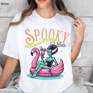 May include: White t-shirt with a colorful graphic. The graphic features a cartoon illustration of a green-skinned woman in a black swimsuit, sitting on a pink flamingo float, holding a cocktail. Text reads "Spooky Summerween Vibes".