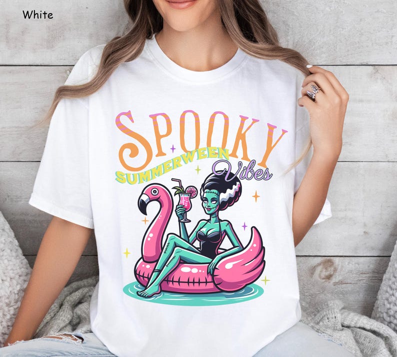 May include: White t-shirt with a colorful graphic. The graphic features a cartoon illustration of a green-skinned woman in a black swimsuit, sitting on a pink flamingo float, holding a cocktail. Text reads "Spooky Summerween Vibes".