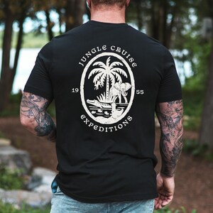 May include: Black t-shirt with a white oval graphic featuring a palm tree, elephant, crocodile, and the words "Jungle Cruise Expeditions 1955". The design is distressed for a vintage look.