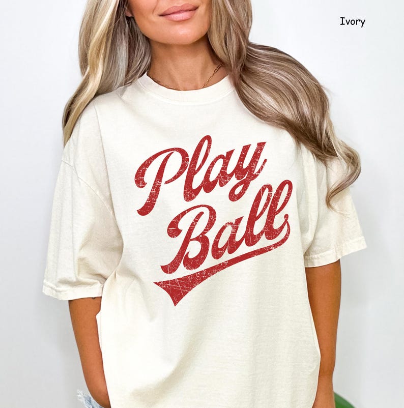 K&ouml;nnte beinhalten: Ein cremefarbenes T-Shirt mit dem Schriftzug "Play Ball" in rot abgenutzten Lettern. Das Shirt hat eine lockere Passform und kurze &Auml;rmel.