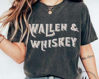 Könnte beinhalten: Ein dunkelgraues T-Shirt mit dem Aufdruck "Wallen & Whiskey" in Weiß. Das Shirt wird von einer Person mit langen blonden Haaren und einer blauen Jeans getragen.