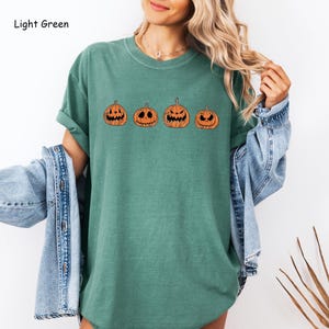 May include: A light green t-shirt featuring a row of five orange pumpkins with different facial expressions. The pumpkins are outlined in black.