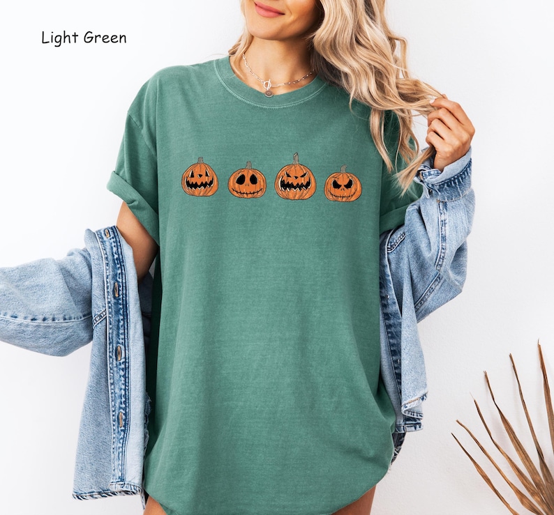 May include: A light green t-shirt featuring a row of five orange pumpkins with different facial expressions. The pumpkins are outlined in black.