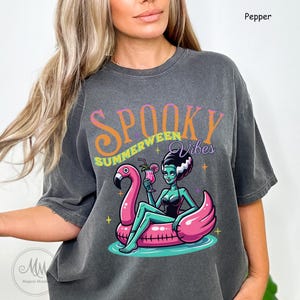 May include: Dark gray t-shirt with the words "Spooky Summerween Vibes" in orange and purple. The graphic features a cartoon Bride of Frankenstein character on a pink flamingo float, holding a cocktail. The shirt is a relaxed fit.