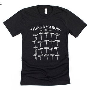May include: A black heather grey t-shirt with a white graphic of the text "Thingamabobs 20" and a collection of 20 different vintage corkscrews.