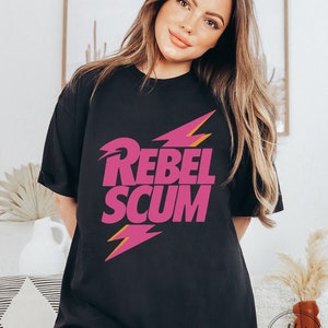 May include: A black short-sleeved t-shirt featuring the text REBEL SCUM in bold pink capital letters. The design includes two stylized pink lightning bolts with orange outlines positioned above and below the text on the front of the shirt.
