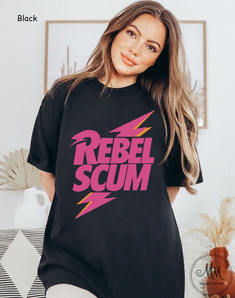 May include: A black short-sleeved t-shirt featuring the text REBEL SCUM in bold pink capital letters. The design includes two stylized pink lightning bolts with orange outlines positioned above and below the text on the front of the shirt.