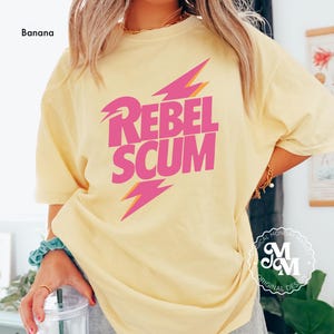 May include: A banana yellow short-sleeve t-shirt featuring the text REBEL SCUM in bold pink letters. The design includes two stylized pink lightning bolts with orange outlines positioned above and below the text on the front of the shirt.