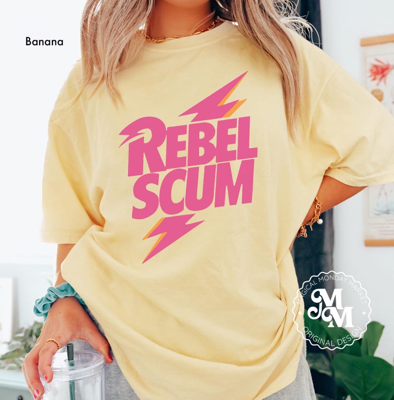 May include: A banana yellow short-sleeve t-shirt featuring the text REBEL SCUM in bold pink letters. The design includes two stylized pink lightning bolts with orange outlines positioned above and below the text on the front of the shirt.