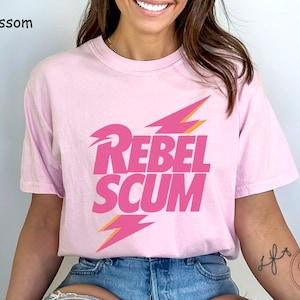 May include: A light pink short-sleeve t-shirt featuring the text REBEL SCUM in bold pink letters. The design includes two stylized lightning bolt graphics in pink and orange, positioned above and below the text. The shirt has a crew neckline.