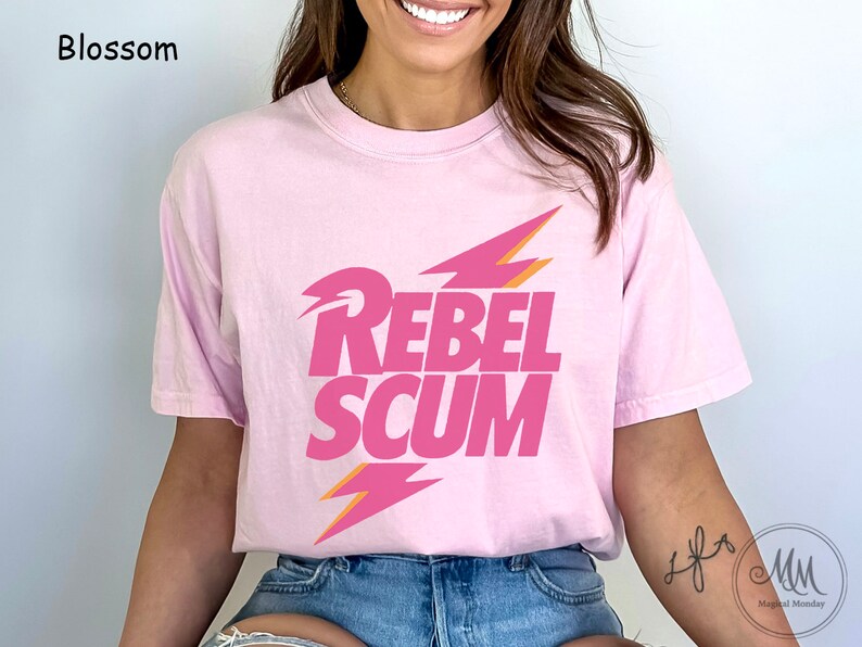 May include: A light pink short-sleeve t-shirt featuring the text REBEL SCUM in bold pink letters. The design includes two stylized lightning bolt graphics in pink and orange, positioned above and below the text. The shirt has a crew neckline.