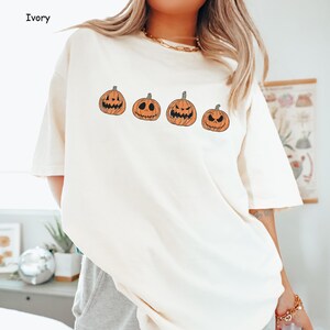 May include: A white t-shirt featuring a graphic of four orange pumpkins with varying facial expressions. The pumpkins are positioned in a line across the front of the shirt.