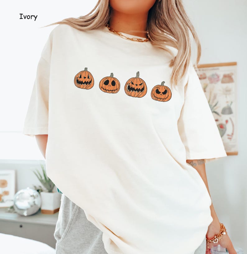 May include: A white t-shirt featuring a graphic of four orange pumpkins with varying facial expressions. The pumpkins are positioned in a line across the front of the shirt.