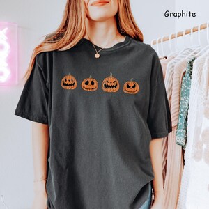 May include: A black t-shirt featuring a row of four orange pumpkins with different facial expressions. The pumpkins are outlined in black. The text "Graphite" is in the top right corner of the image.