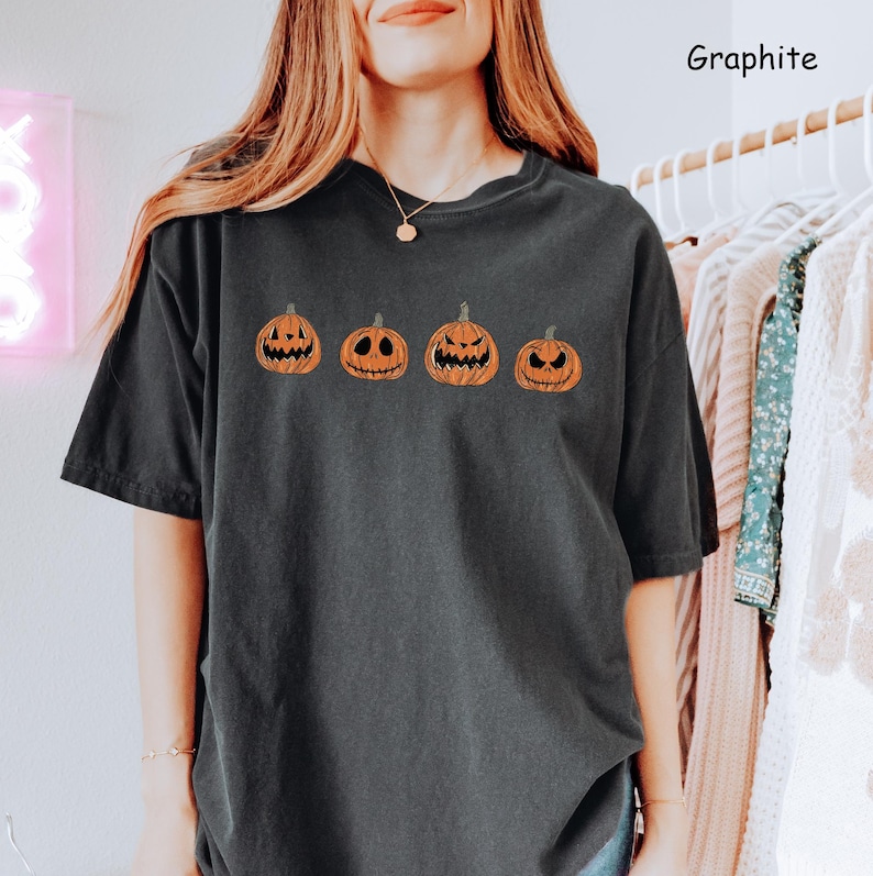 May include: A black t-shirt featuring a row of four orange pumpkins with different facial expressions. The pumpkins are outlined in black. The text "Graphite" is in the top right corner of the image.