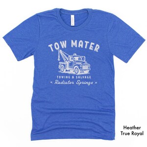 Pu&ograve; includere: T-shirt blu melange con grafica bianca di un carro attrezzi. Il testo sopra il camion recita "TOW MATER" e sotto "TOWING & SALVAGE" e "Radiator Springs". La maglietta &egrave; etichettata "Heather True Royal".