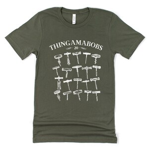 May include: A military green t-shirt with a white graphic of the text "Thingamabobs - 20" and a collection of 20 different types of corkscrews.