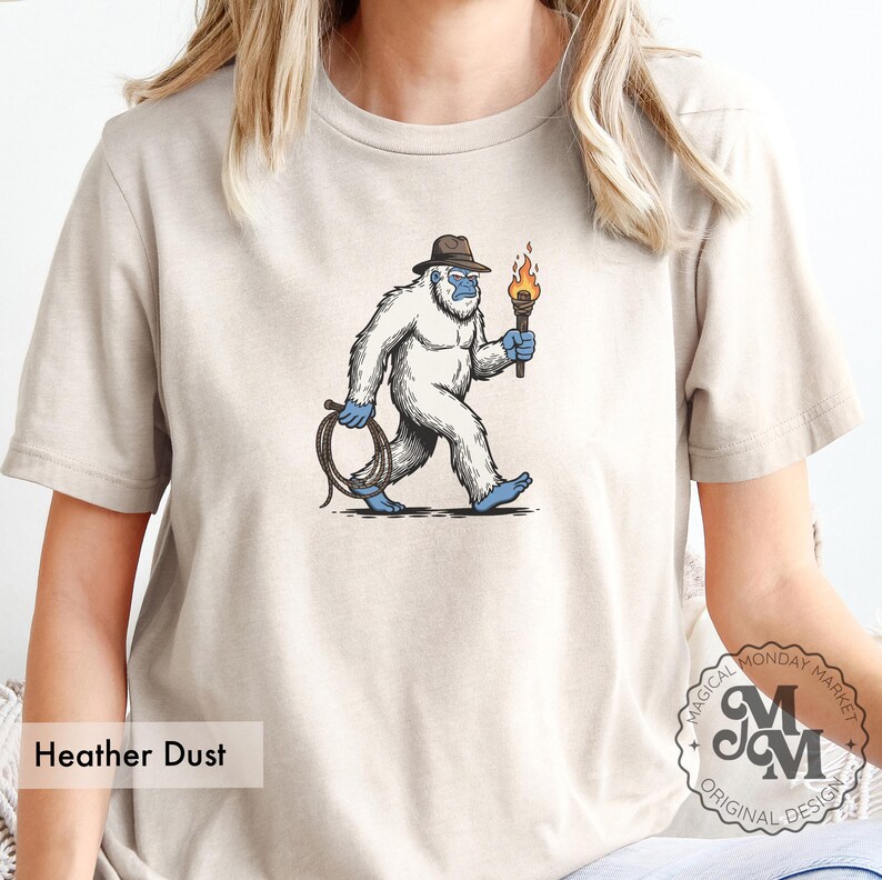 Peut inclure: T-shirt couleur Heather Dust avec une illustration de dessin anim&eacute; d'un y&eacute;ti blanc portant un chapeau marron et tenant une torche et une corde. Le y&eacute;ti a les pieds et les mains bleus. Le t-shirt porte le texte "Heather Dust".