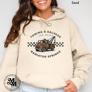 May include: Sand-coloured hoodie with a graphic of a vintage tow truck and the text "Towing & Salvage Tow Mater Radiator Springs." The design includes a checkered pattern and a logo. The hoodie has a drawstring hood and a front pocket.