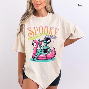 May include: Ivory-colored t-shirt with a graphic of a cartoon woman in a black swimsuit and green skin, floating on a pink flamingo pool float. The text "Spooky Summerween Vibes" is above the image.