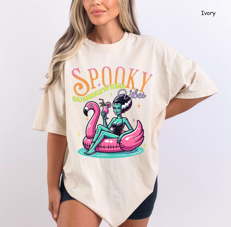 May include: Ivory-colored t-shirt with a graphic of a cartoon woman in a black swimsuit and green skin, floating on a pink flamingo pool float. The text "Spooky Summerween Vibes" is above the image.