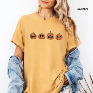 May include: A mustard yellow t-shirt featuring a row of five orange jack-o'-lantern pumpkins with different facial expressions.