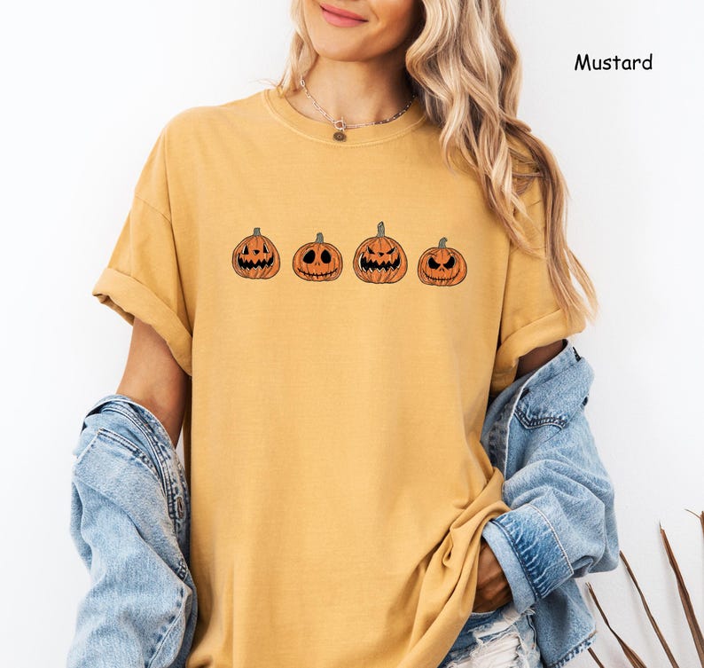 May include: A mustard yellow t-shirt featuring a row of five orange jack-o'-lantern pumpkins with different facial expressions.