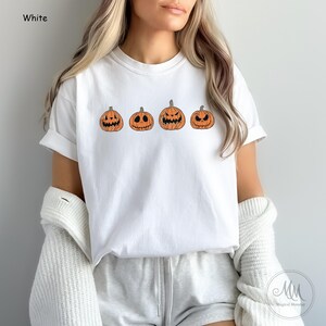 May include: White t-shirt featuring a graphic of four orange pumpkins with different facial expressions. The pumpkins are arranged in a row from left to right.
