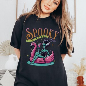 May include: Black t-shirt featuring a cartoon illustration of a green-skinned woman with white hair, lounging on a pink flamingo float in water. The shirt has the words "Spooky Summerween Vibes" in a colorful, stylized font.