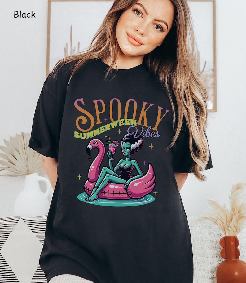 May include: Black t-shirt featuring a cartoon illustration of a green-skinned woman with white hair, lounging on a pink flamingo float in water. The shirt has the words "Spooky Summerween Vibes" in a colorful, stylized font.