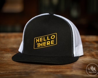 May include: A black and white trucker hat with a white mesh back. The front of the hat has a black embroidered rectangle with the words "HELLO THERE" in yellow.