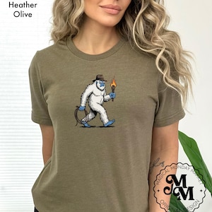 Peut inclure: T-shirt vert olive chin&eacute; avec un dessin de y&eacute;ti portant un chapeau et tenant une torche et une corde. Le y&eacute;ti est blanc avec des accents bleus. Le t-shirt porte le texte "Magical Monday Market Original Design" en bas &agrave; droite.