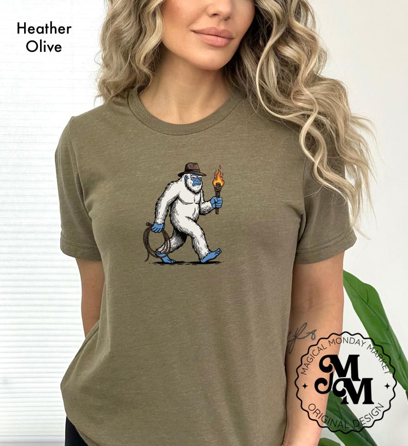Peut inclure: T-shirt vert olive chin&eacute; avec un dessin de y&eacute;ti portant un chapeau et tenant une torche et une corde. Le y&eacute;ti est blanc avec des accents bleus. Le t-shirt porte le texte "Magical Monday Market Original Design" en bas &agrave; droite.