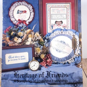 May include: A collection of framed cross-stitch samplers and a pocket watch are displayed on a blue surface. The samplers feature floral and script designs, with one displaying the text "God bless you, precious friend." The title "Heritage of Friends" is visible.