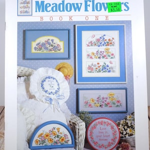 May include: A book cover for a cross-stitch pattern book titled "Meadow Flowers Book One" by Karen Avery. The cover features a white wicker chair with a white pillow and a variety of cross-stitch projects displayed on the chair and the wall behind it. The projects include a pillow, a wall hanging, a round hoop, and a half-round hoop. The projects feature floral designs and the text "Love lives in your heart Forever".