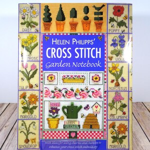 Puede incluir: Un cuaderno de jardín de punto de cruz con un fondo amarillo y una variedad de flores bordadas, macetas y elementos de jardín. La portada presenta el título "Helen Philipps' Cross Stitch Garden Notebook".