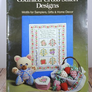 May include: A book titled "Counted Cross-Stitch Designs" with a blue background and a cross-stitch design of a floral sampler. The book features a teddy bear, a heart-shaped box, and a basket of strawberries.