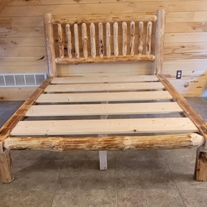 May include: A rustic wooden bed frame with a headboard featuring vertical log accents. The bed frame is constructed from natural wood, showcasing the knots and textures of the material. The bed frame has a slatted base.