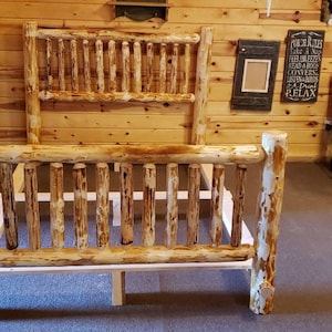 May include: A rustic wooden bed frame with a whitewashed finish. The headboard and footboard are made of thick, natural logs with a rough texture. The frame has a simple design with vertical slats.