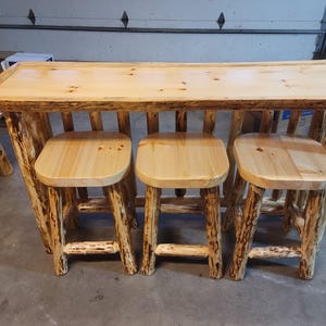 May include: A rustic wooden bar with three matching stools. The bar and stools are made from light-coloured wood, with a natural, unfinished appearance. The bar has a rectangular top and the stools have square seats.