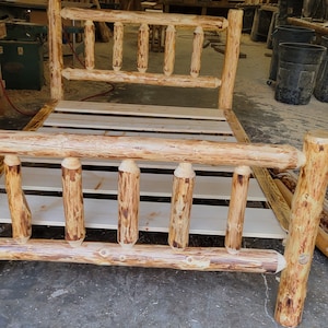 May include: A rustic wooden bed frame made from natural logs with a light brown finish. The bed frame has a slatted headboard and footboard, and the slats are made from unfinished wood. The bed frame is sitting on a concrete floor in a workshop.
