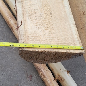 May include: A light-colored wooden plank with a yellow measuring tape showing approximately 11 inches. The wood has visible grain and texture. Two wooden poles are in the background.