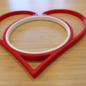 May include: A red heart-shaped embroidery hoop frame with a white circular inner hoop. The red outer frame forms a distinct heart outline, while the white inner ring is positioned centrally to hold fabric securely within the heart-shaped structure.